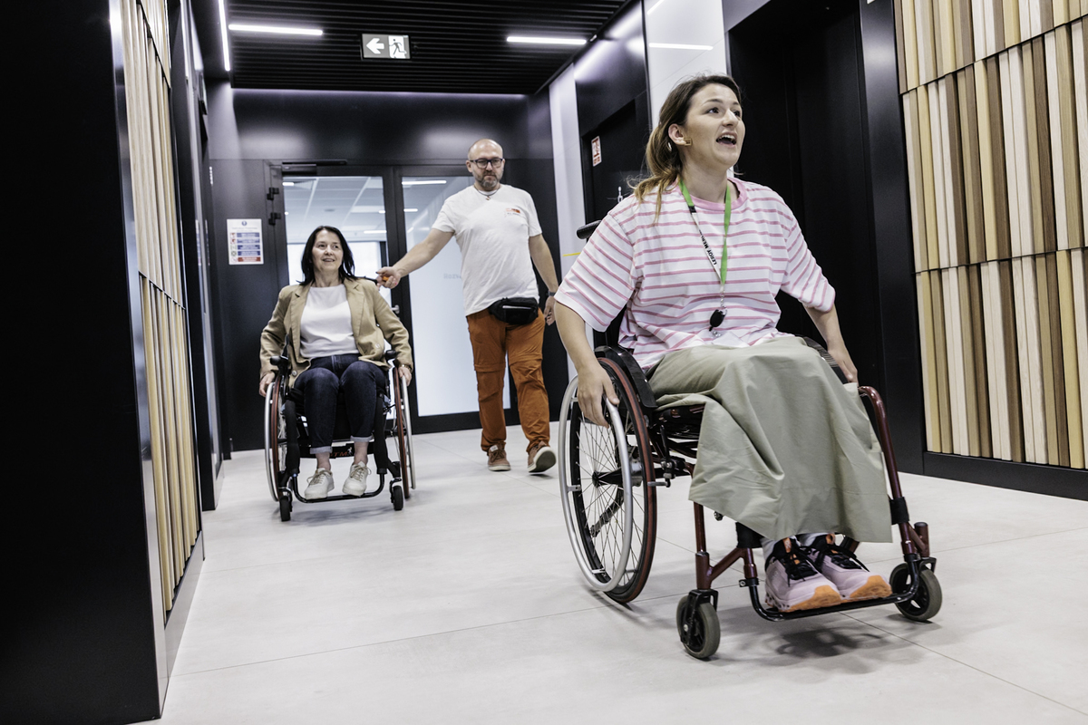 Two people ride independently in active wheelchairs in the office corridor of Leroy Merlin Poland. In the background, a man from the Foundation's team walks behind them, supporting the exercise.